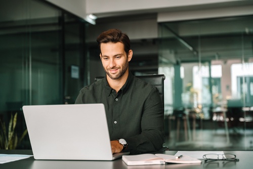 Professional seated at desk using laptop with notebook and eyeglasses nearby