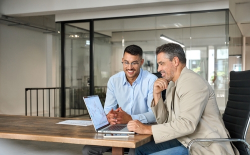 Two businessmen reviewing laptop screen at office table with printed financial charts