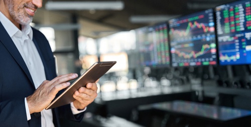 businessman holding tablet in trading room with stock market screens