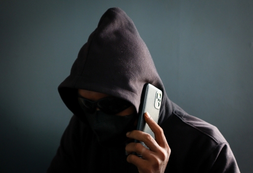 masked hooded figure holding smartphone to ear depicting phone scammer